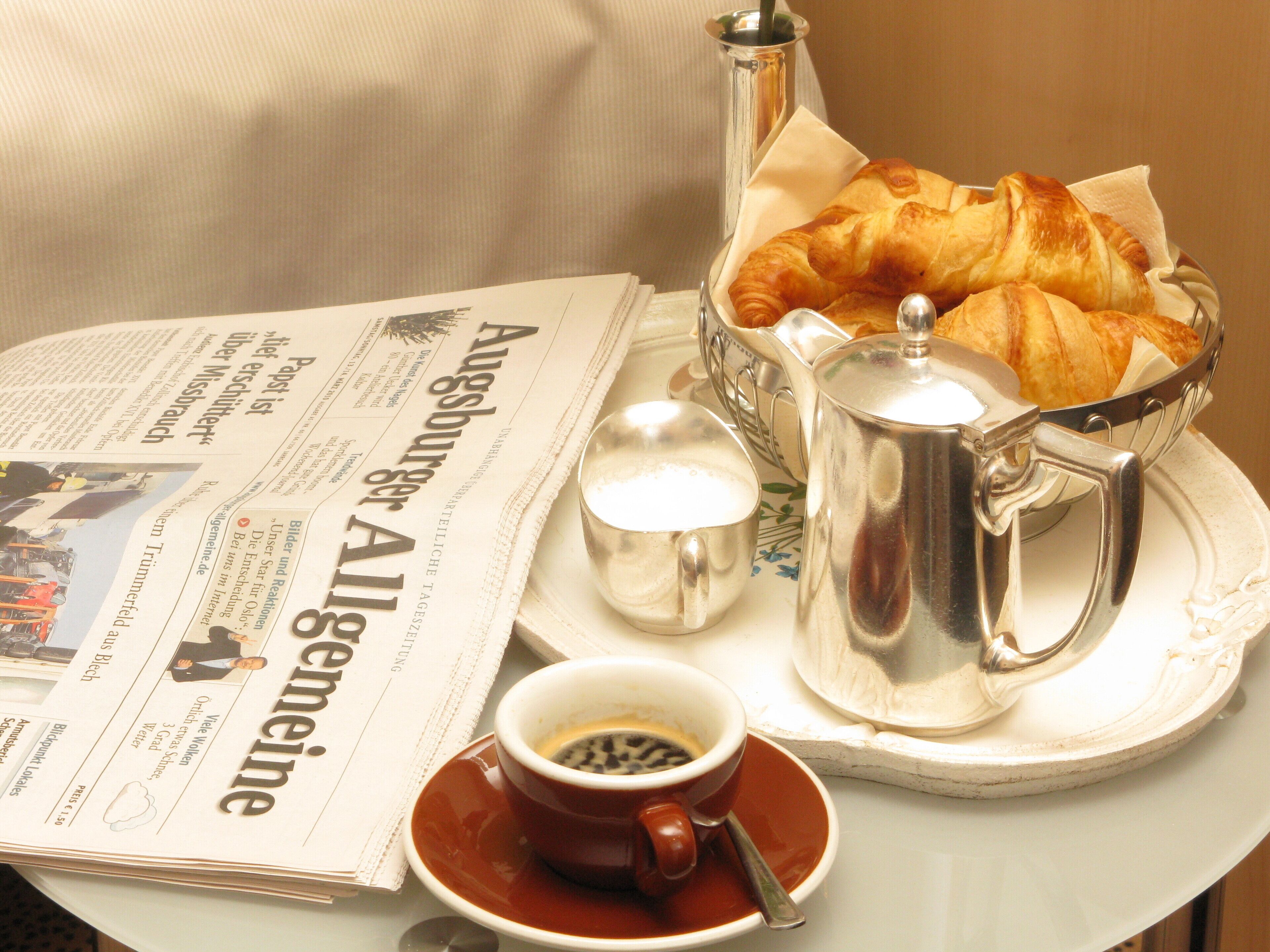 A table with a newspaper, a croissant basket, a coffee pot, and a cup of coffee.