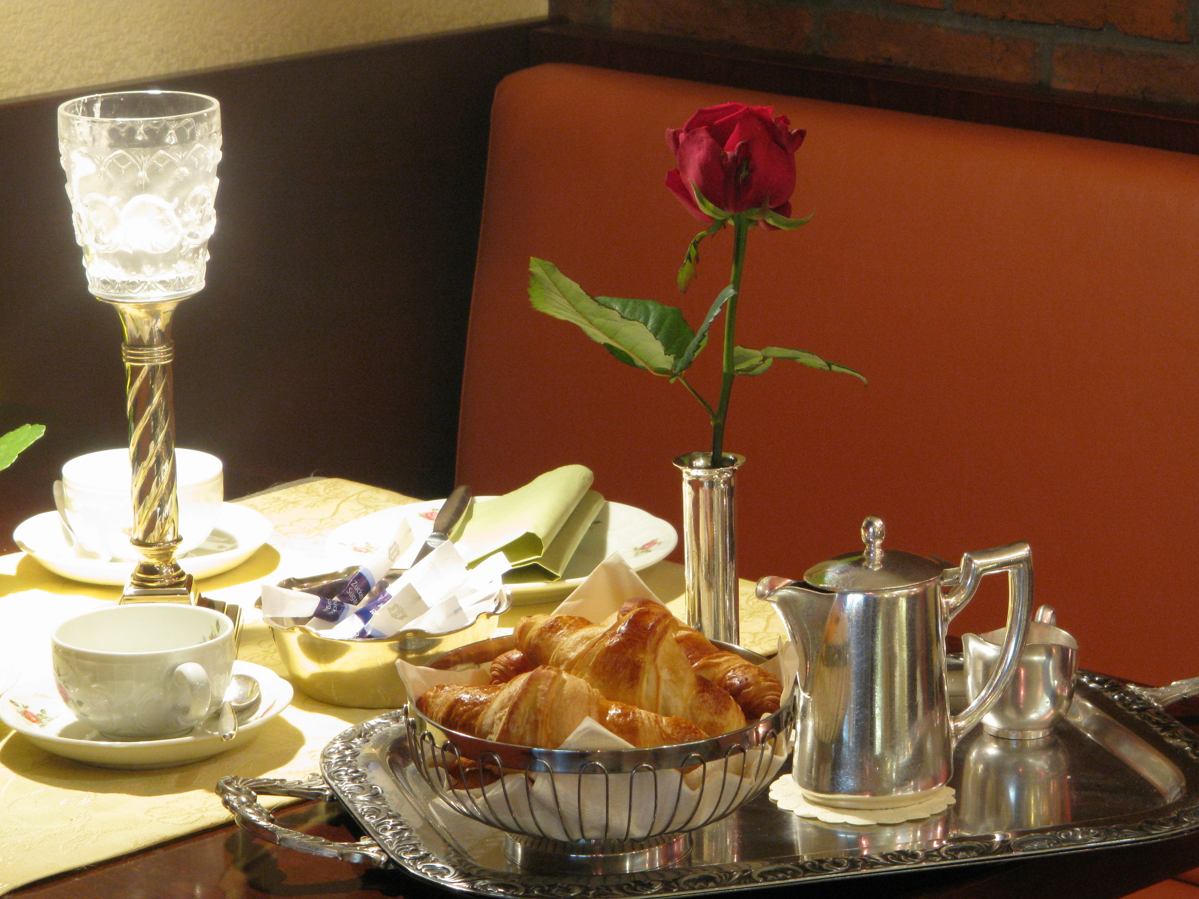 Une table à manger sur laquelle se trouve un plateau contenant des croissants, une théière, un sucrier, un pot à lait et une rose dans un vase.