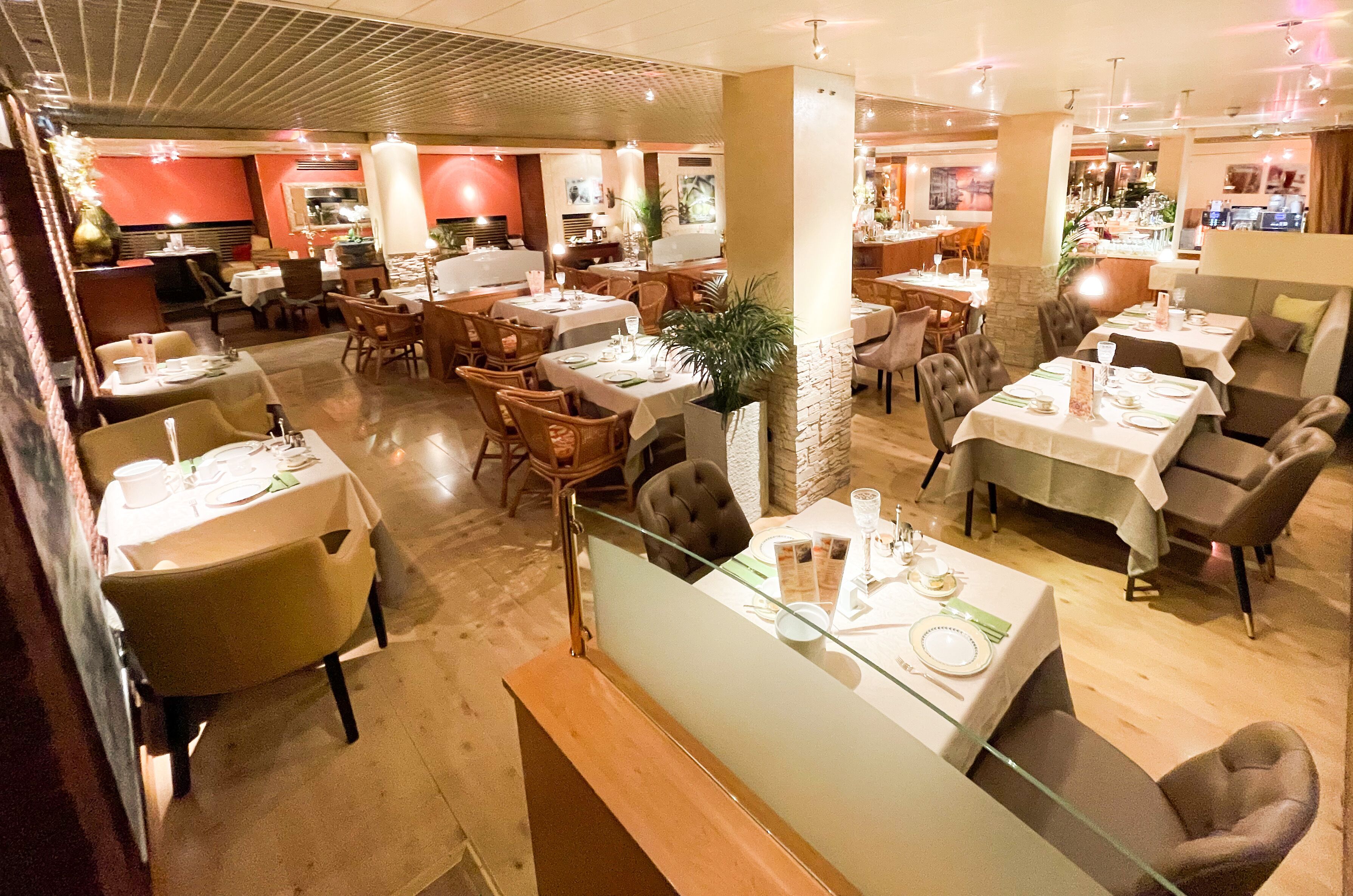 A restaurant with multiple dining areas, including a bar and a seating area with a glass partition. Tables are set with white tablecloths and silverware.