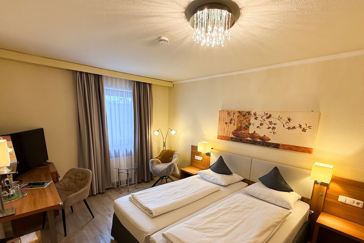A hotel room with two beds, a desk with a computer, a chair, a lamp, and a painting on the wall.