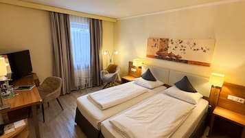 Habitación de hotel con dos camas, un escritorio con computadora, una silla, una lámpara y un cuadro en la pared.