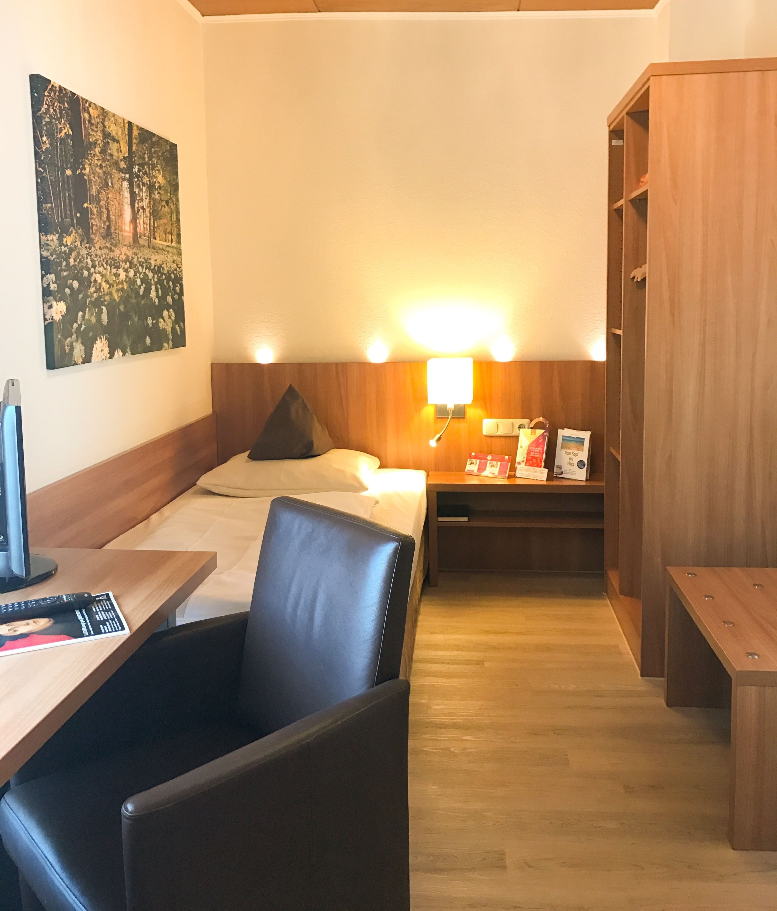 Una habitación de hotel con una cama, un escritorio con silla, una mesita pequeña, una mesita de noche y un armario de madera.