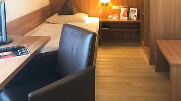 Una habitación de hotel con una cama, un escritorio con silla, una mesita pequeña, una mesita de noche y un armario de madera.