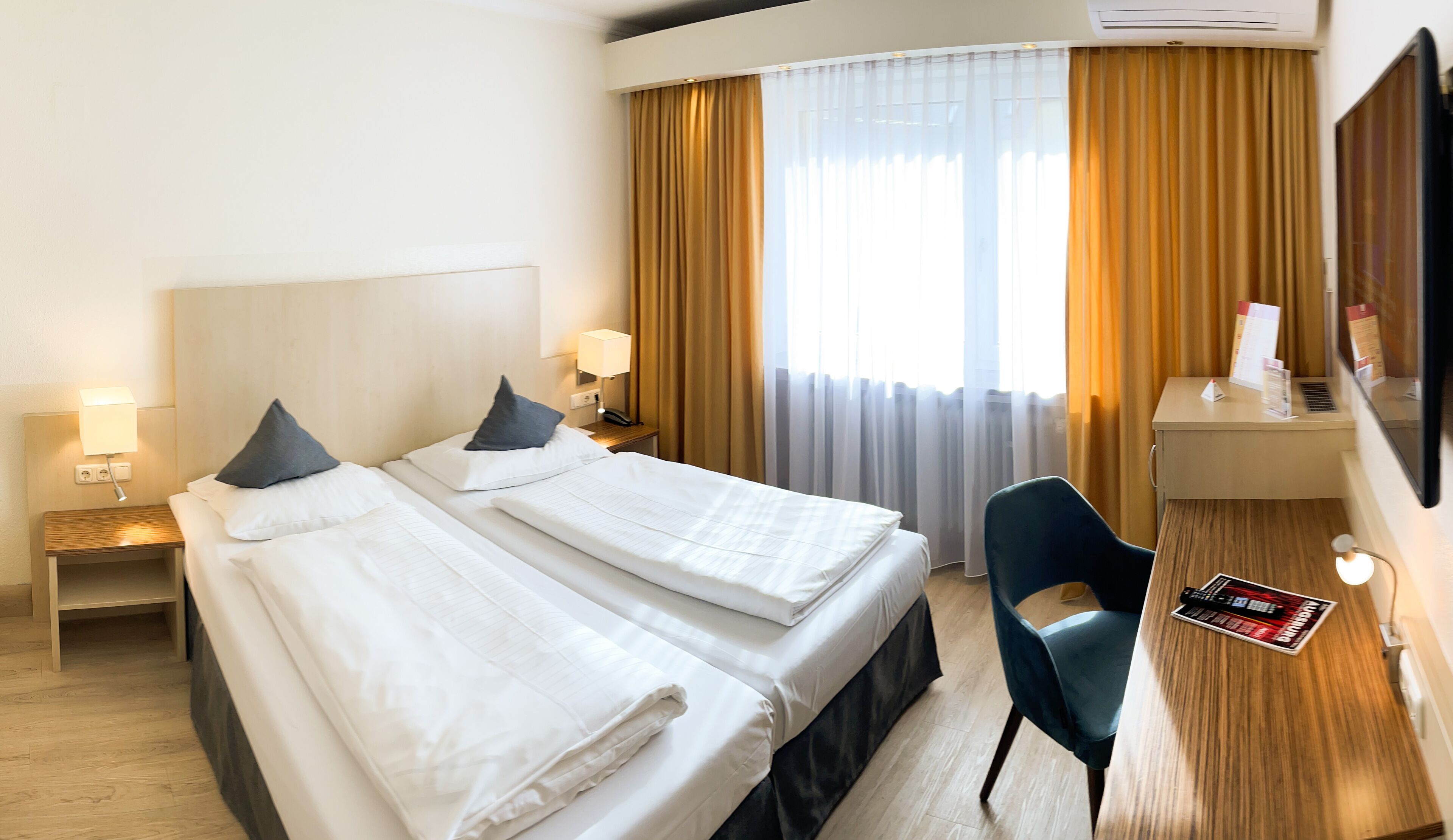 Ein Hotelzimmer mit zwei Betten, einem Schreibtisch, einem Stuhl und einem großen Fenster mit Vorhängen.
