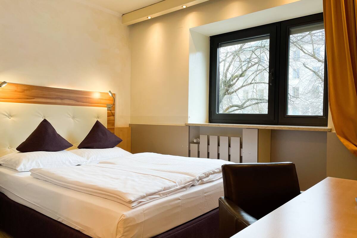 A hotel room with a bed, a window with a view of bare trees, a table, and a chair.