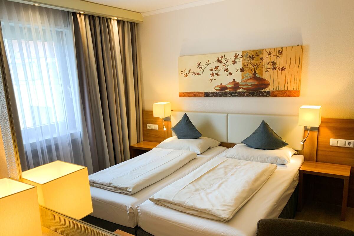 A hotel room with two beds, a painting on the wall, and two bedside lamps.