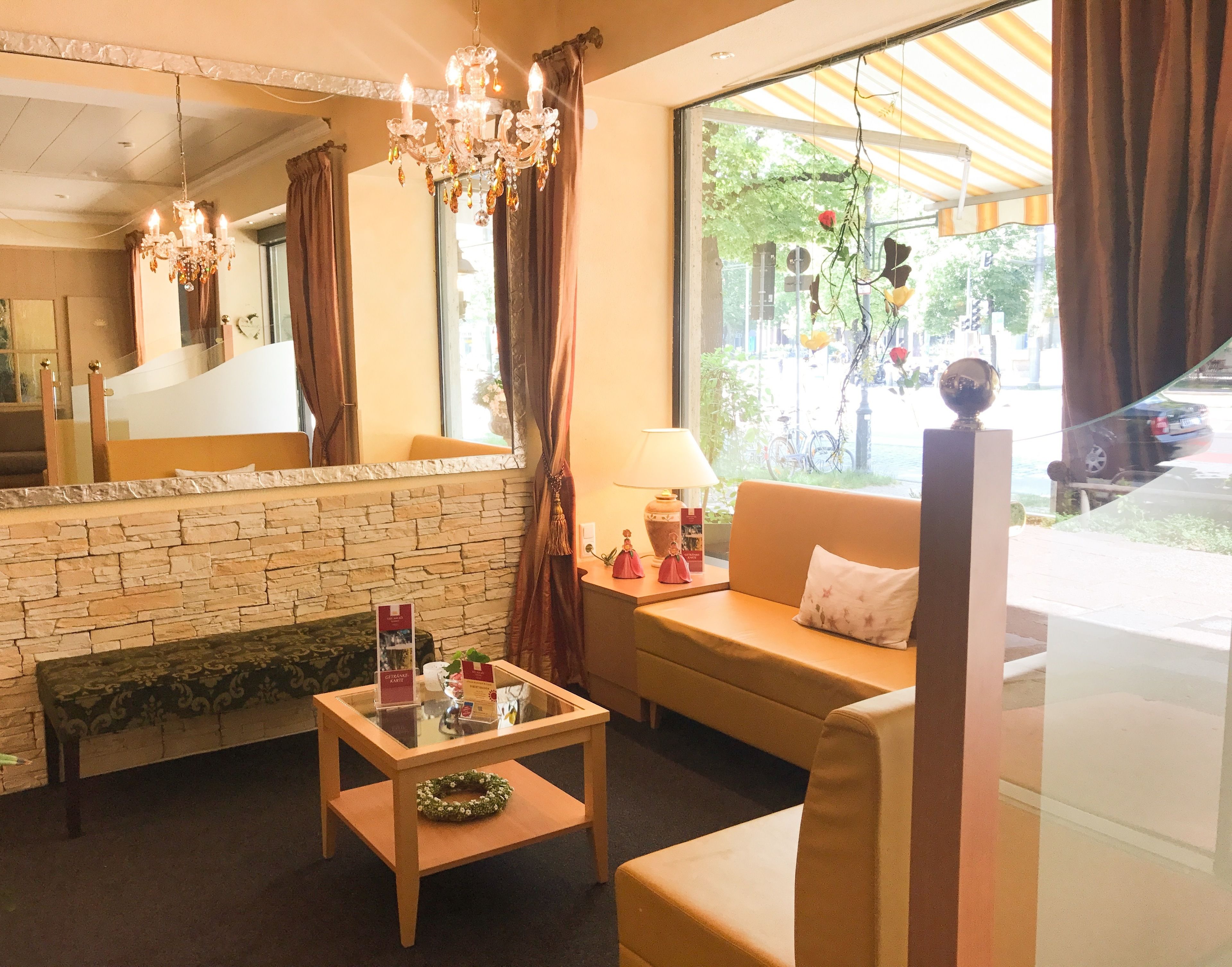 A hotel room with a stone wall, a wooden bench, a glass table, a sofa, a chandelier, and a view of the street through large windows.