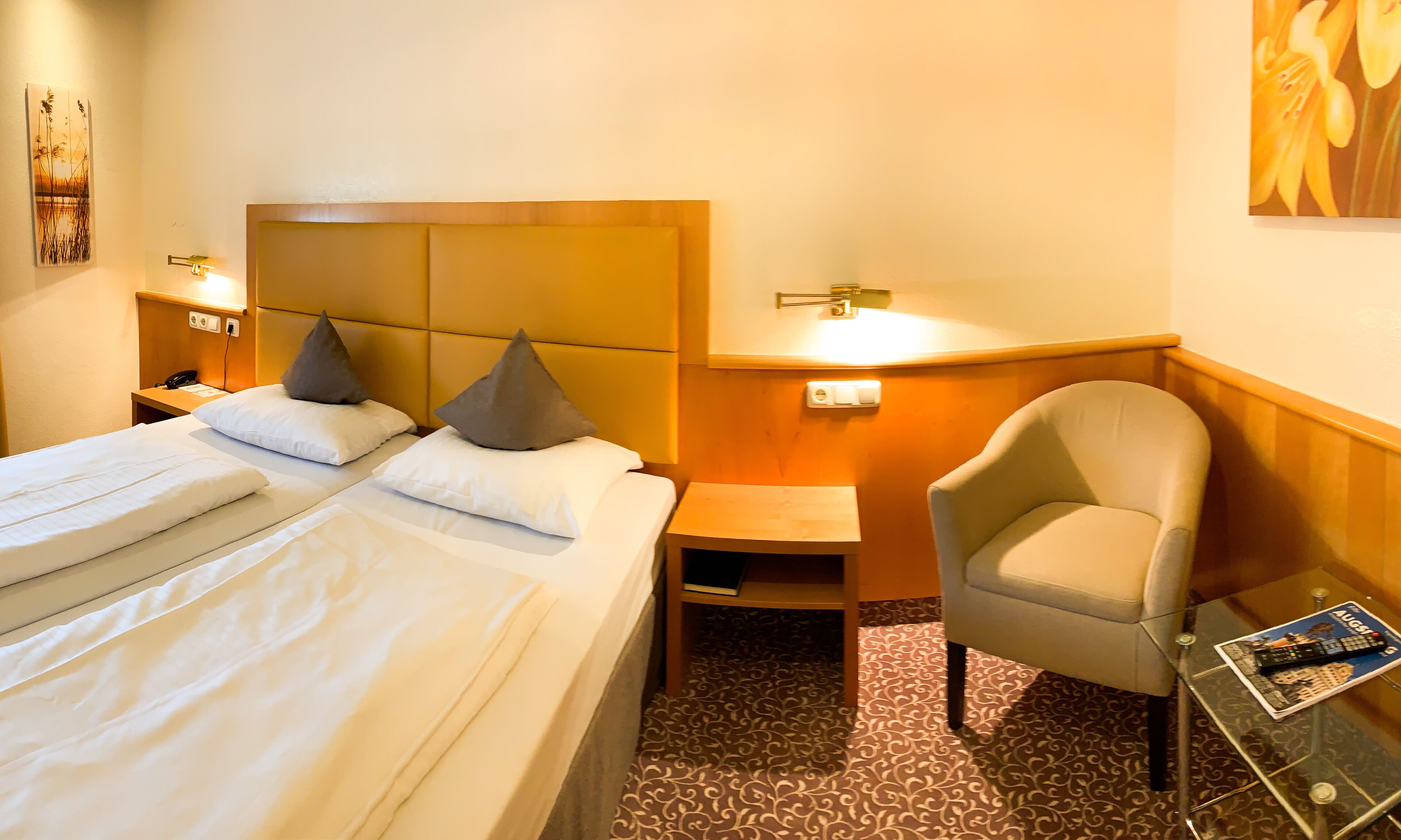 Quarto de hotel com duas camas, uma poltrona, mesa de cabeceira e uma pequena mesa com uma revista.