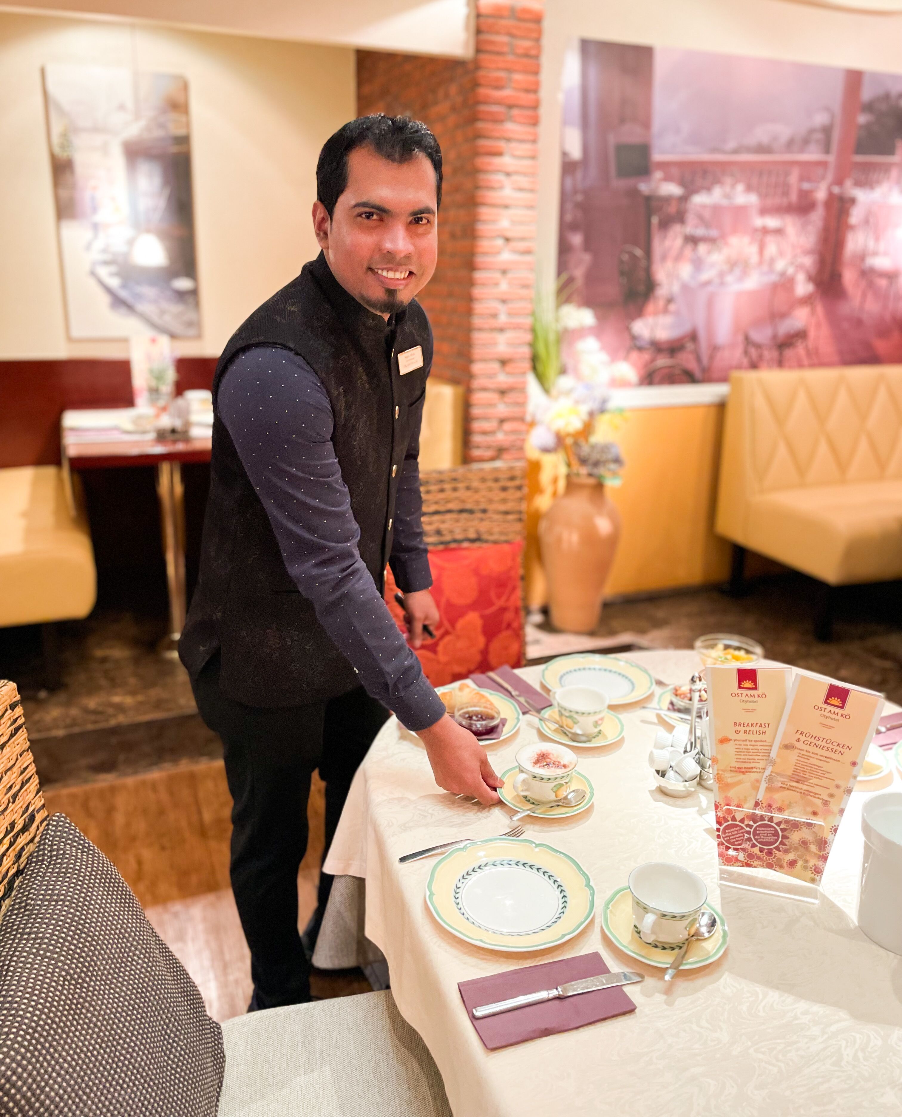 Una persona preparando una mesa con platos, vasos y cubiertos en un restaurante con una pared de ladrillo y un cartel grande al fondo.