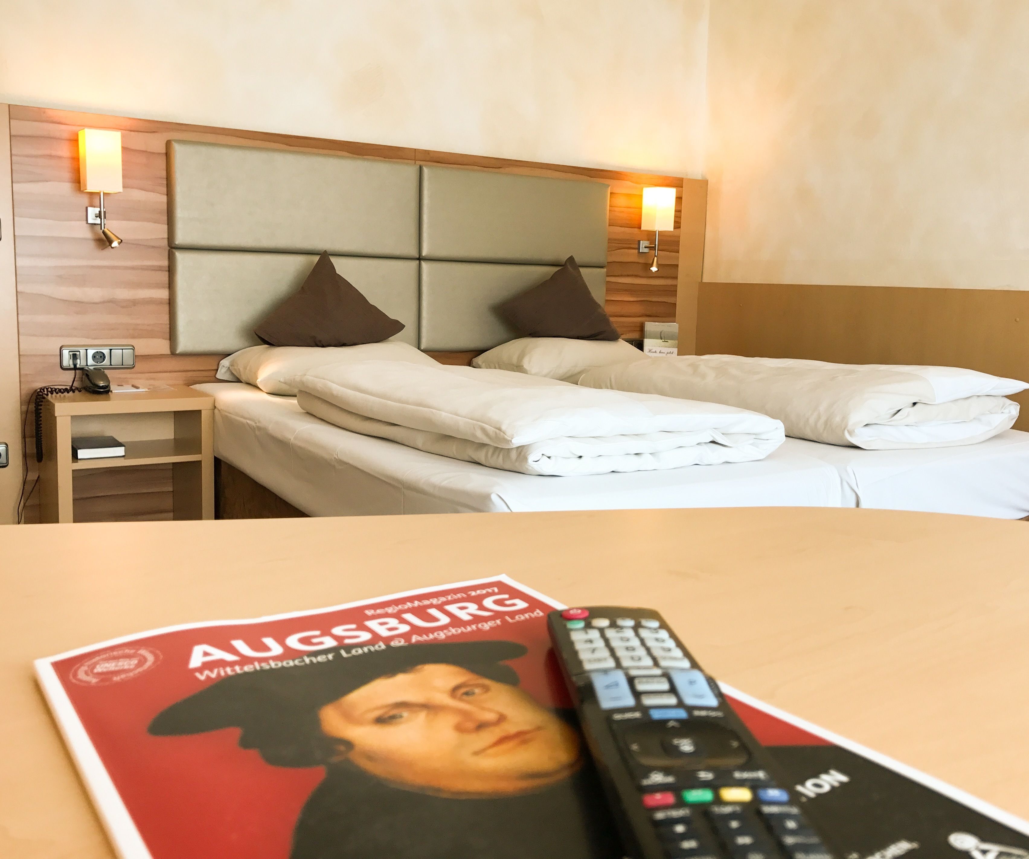 Habitación de hotel con una cama, lámparas de noche, una mesita de noche con un teléfono y una revista sobre la mesa.
