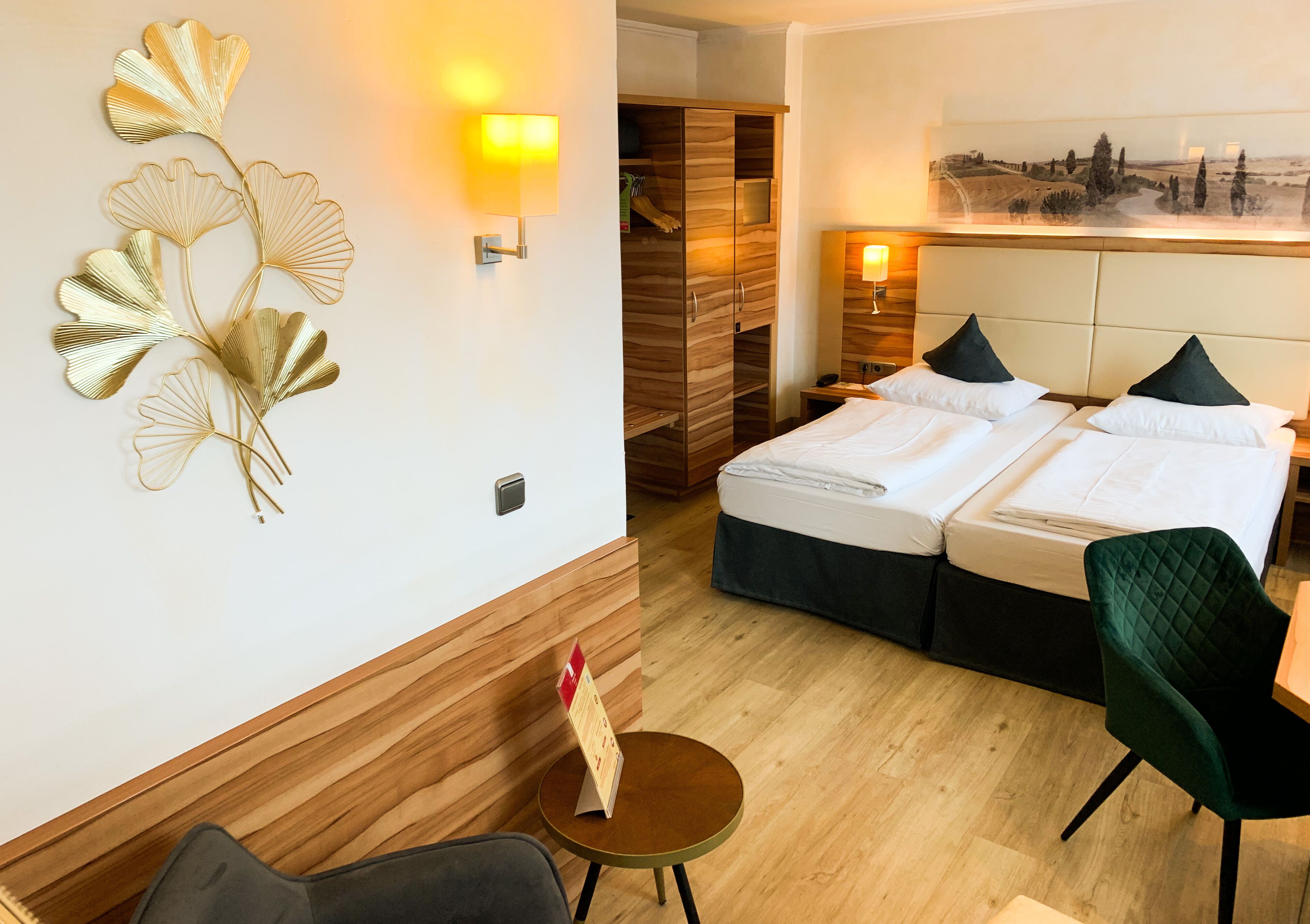 A hotel room with a bed, two bedside tables, a chair, a small round table with a sign, and a wall-mounted lamp.