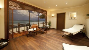 Una habitación con piso de madera, balcón con vista al mar, una silla reclinable y una mesa con sillas.
