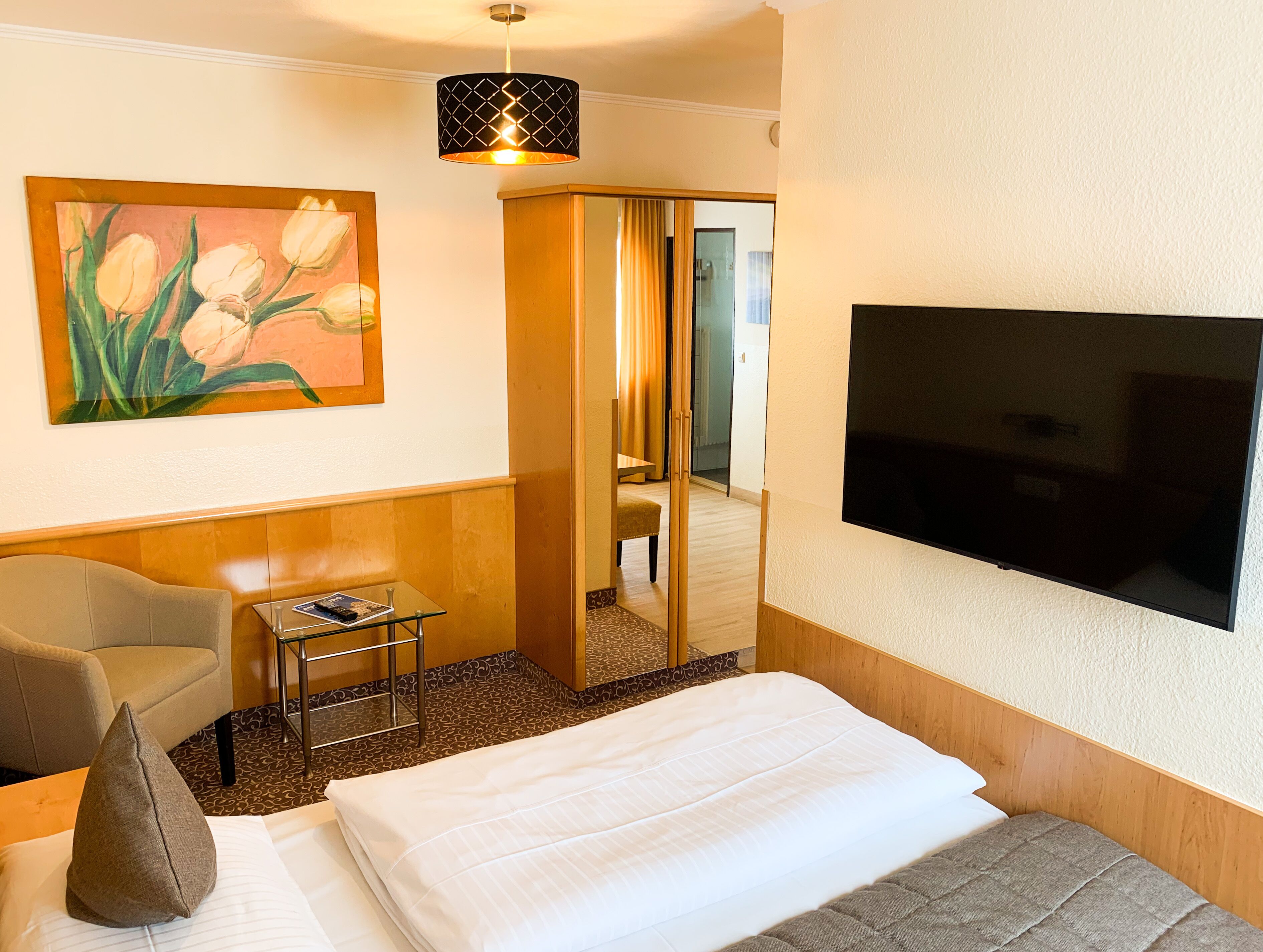 A hotel room with a bed, a chair, a small table, a painting, a TV, and a mirror.