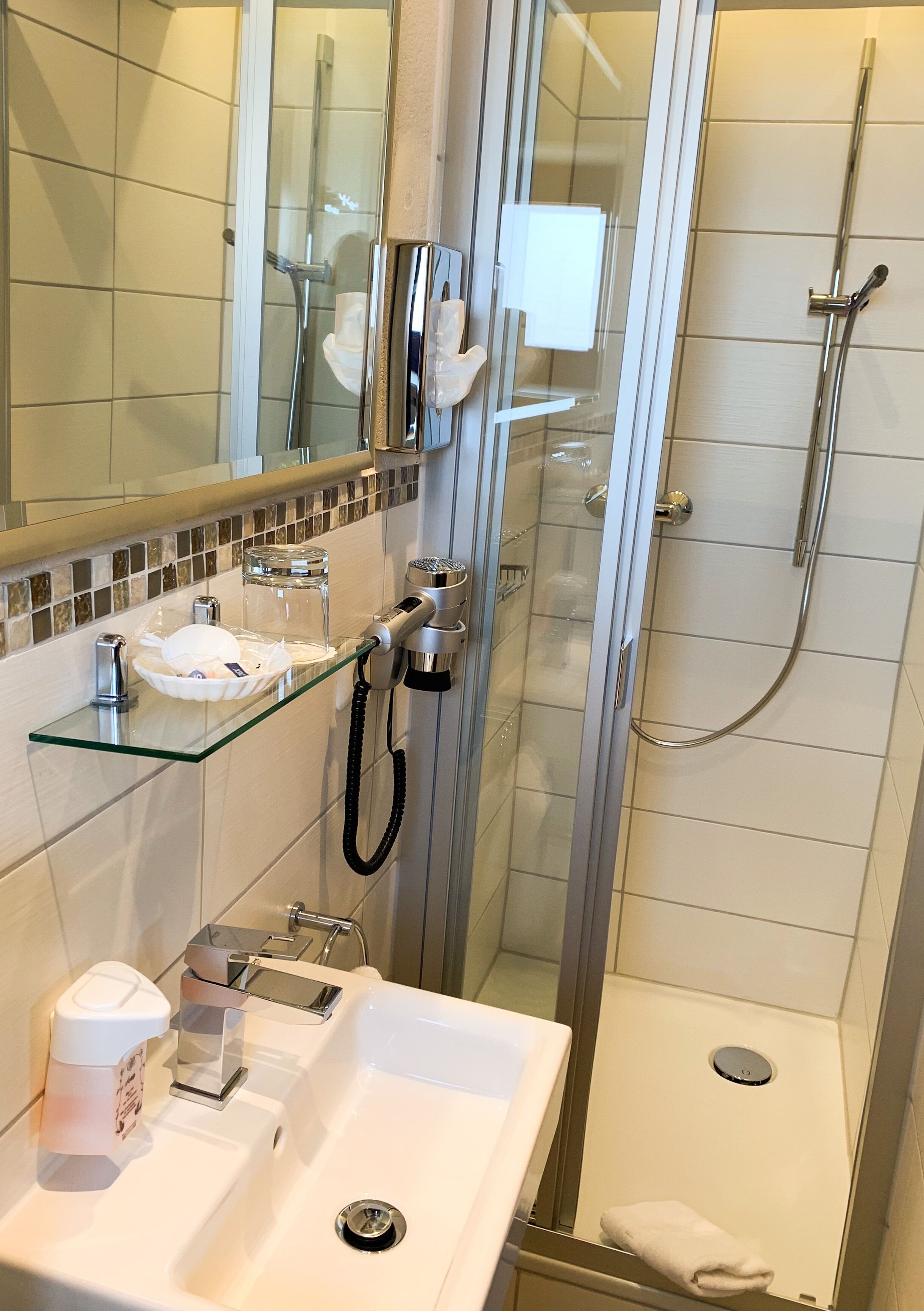 A modern bathroom with a glass shower enclosure, a glass shelf with toiletries, a white sink, and a handheld showerhead.