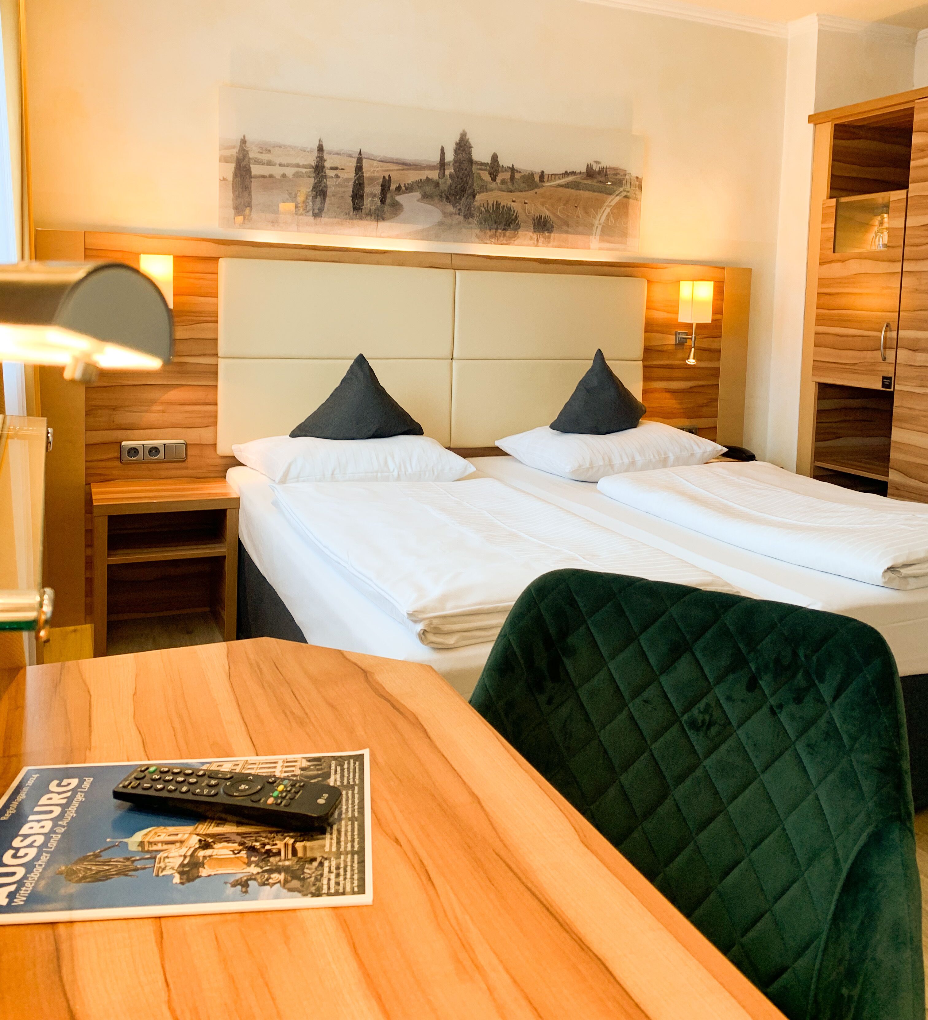 A hotel room with two beds, a wooden table, a green chair, a lamp, and a painting on the wall.