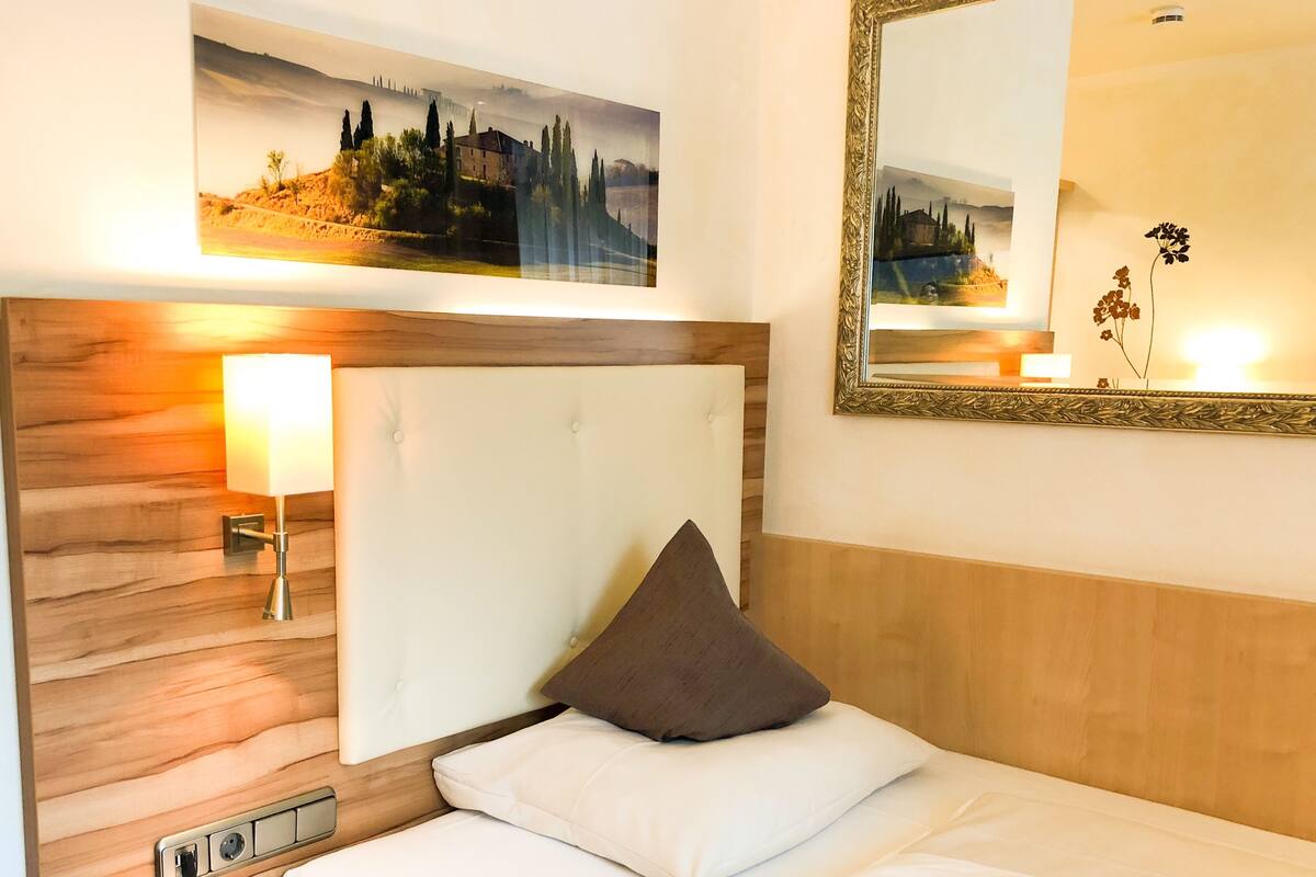 A hotel room with a bed, a bedside lamp, a telephone, a wall-mounted mirror, and a painting above the bed.