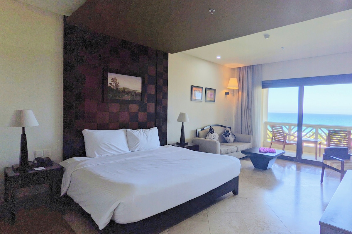 Une chambre d’hôtel avec un grand lit, un canapé, un bureau et un balcon donnant sur la mer.