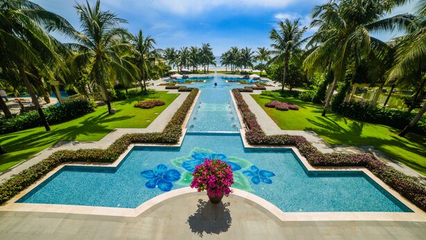 Piscina de resort com um elemento central de água, cercada por palmeiras e gramados bem cuidados.