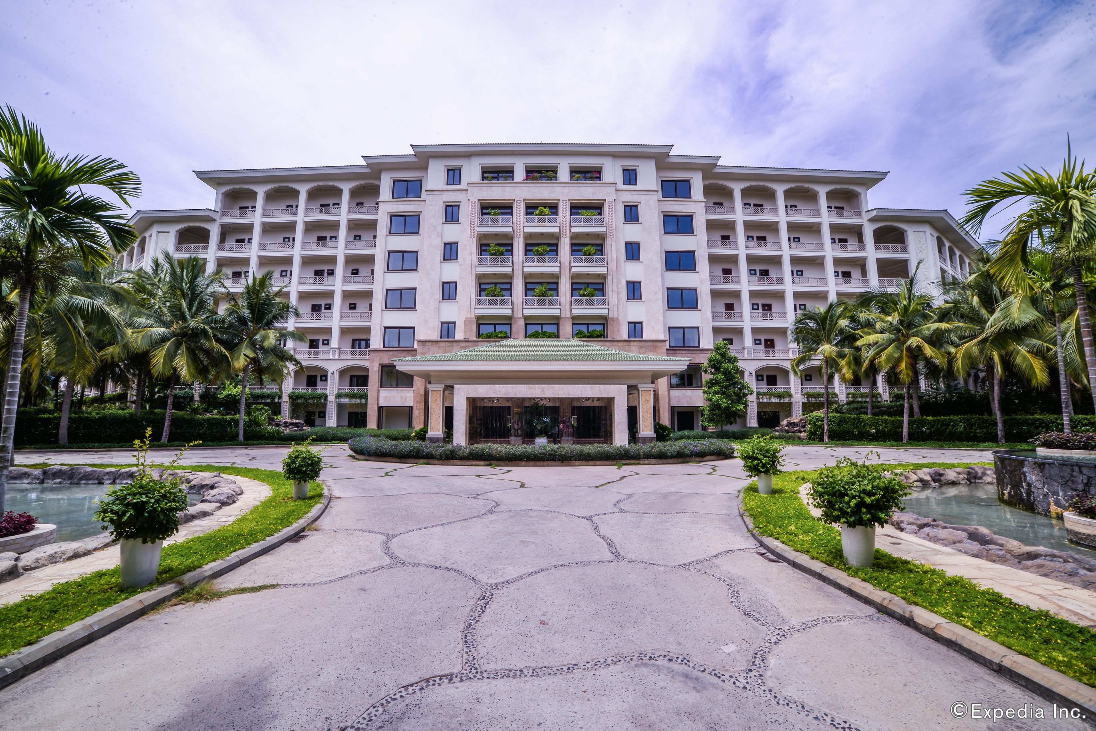 A large, multi-story hotel with a symmetrical facade, a central entrance, and a landscaped courtyard with palm trees and a water feature.