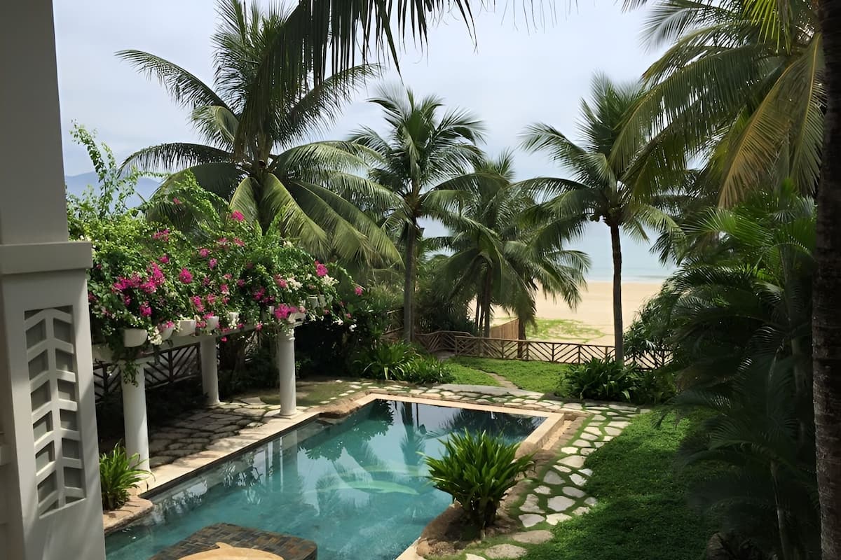 Un espace piscine offrant une vue dégagée sur la plage, entouré de palmiers et d’une clôture blanche.