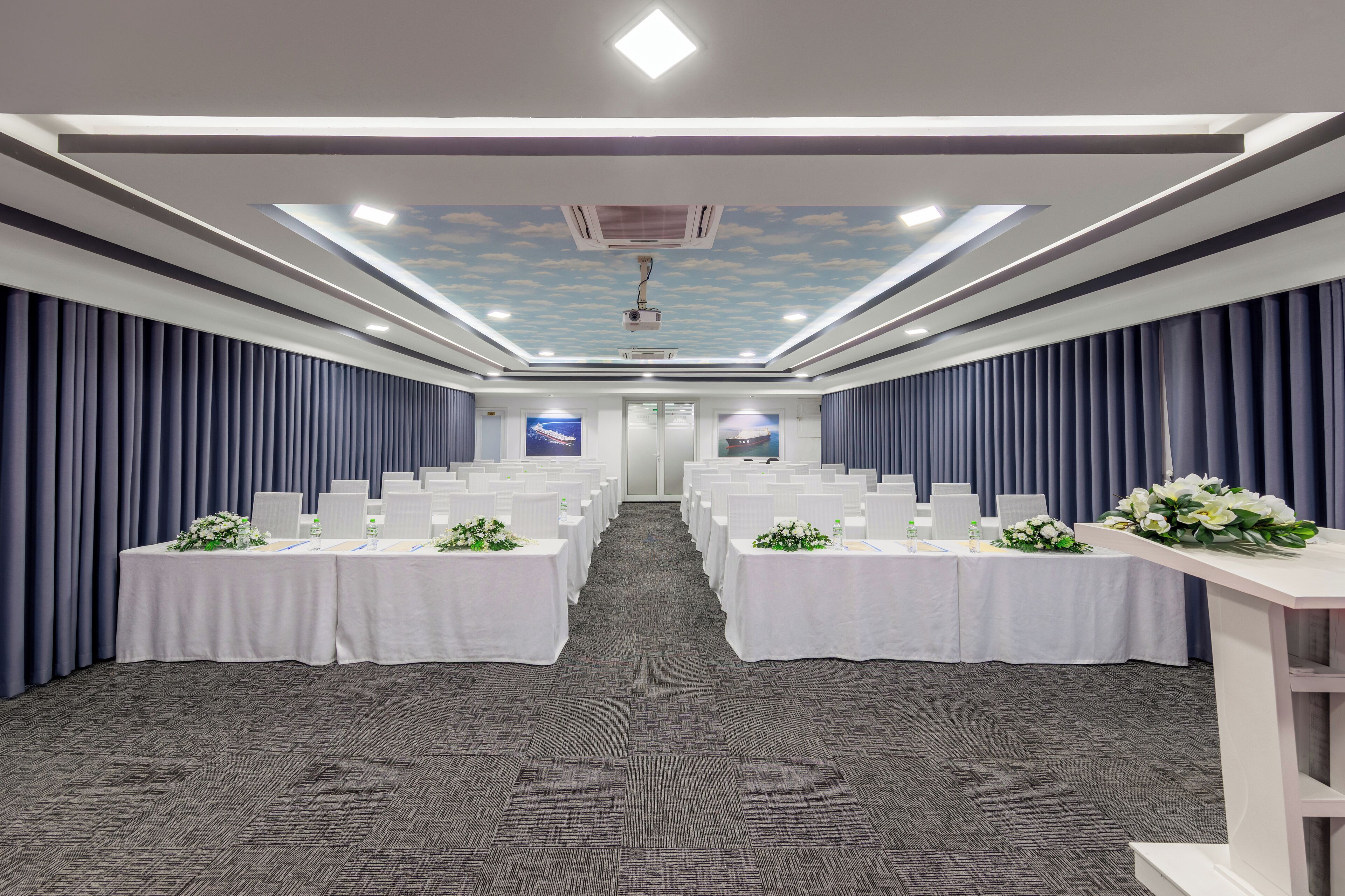 A conference room with rows of tables, a carpeted floor, and a ceiling with a skylight and air conditioning unit.