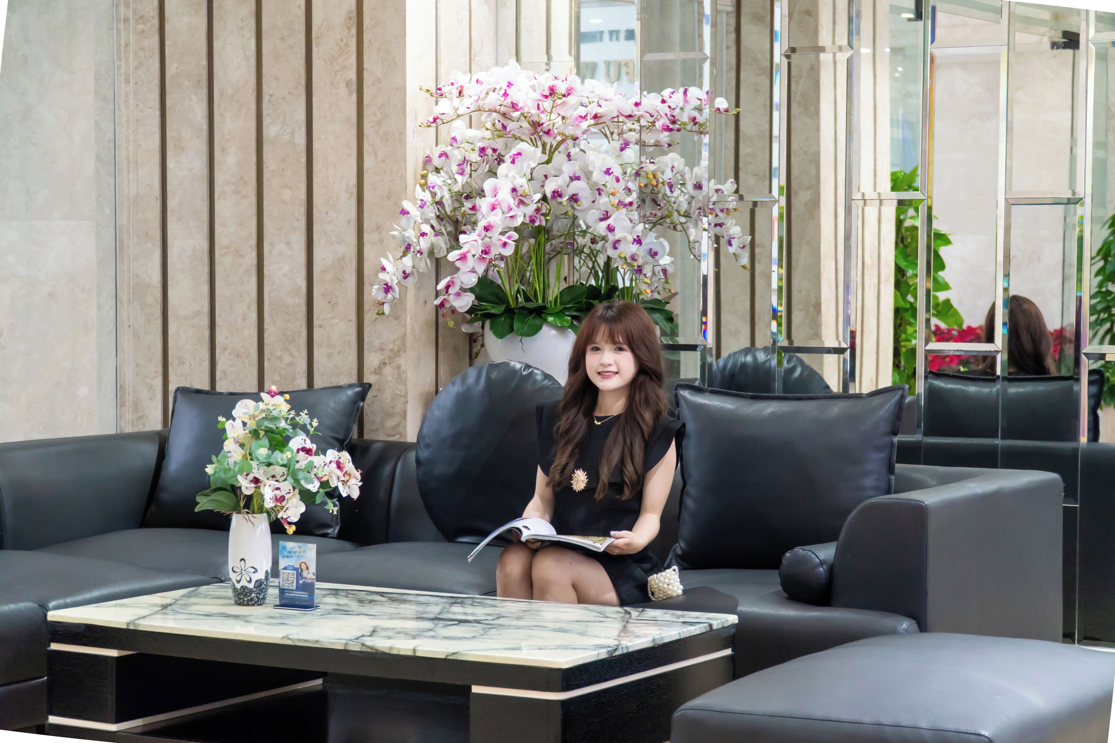 A woman sitting on a black leather sofa with a white vase of flowers and a brochure on a marble coffee table.