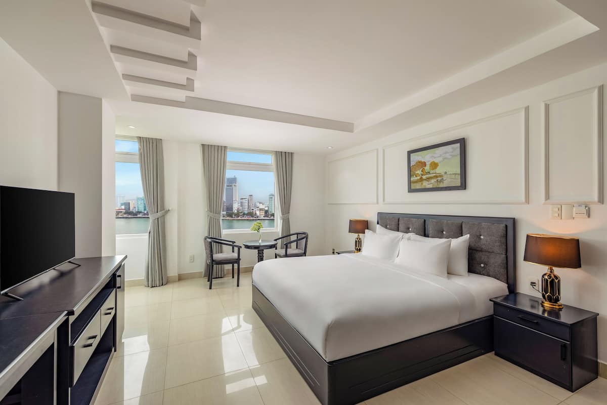 A hotel room with a large bed, a TV, a desk with two chairs, a view of the city, and a cityscape through the window.
