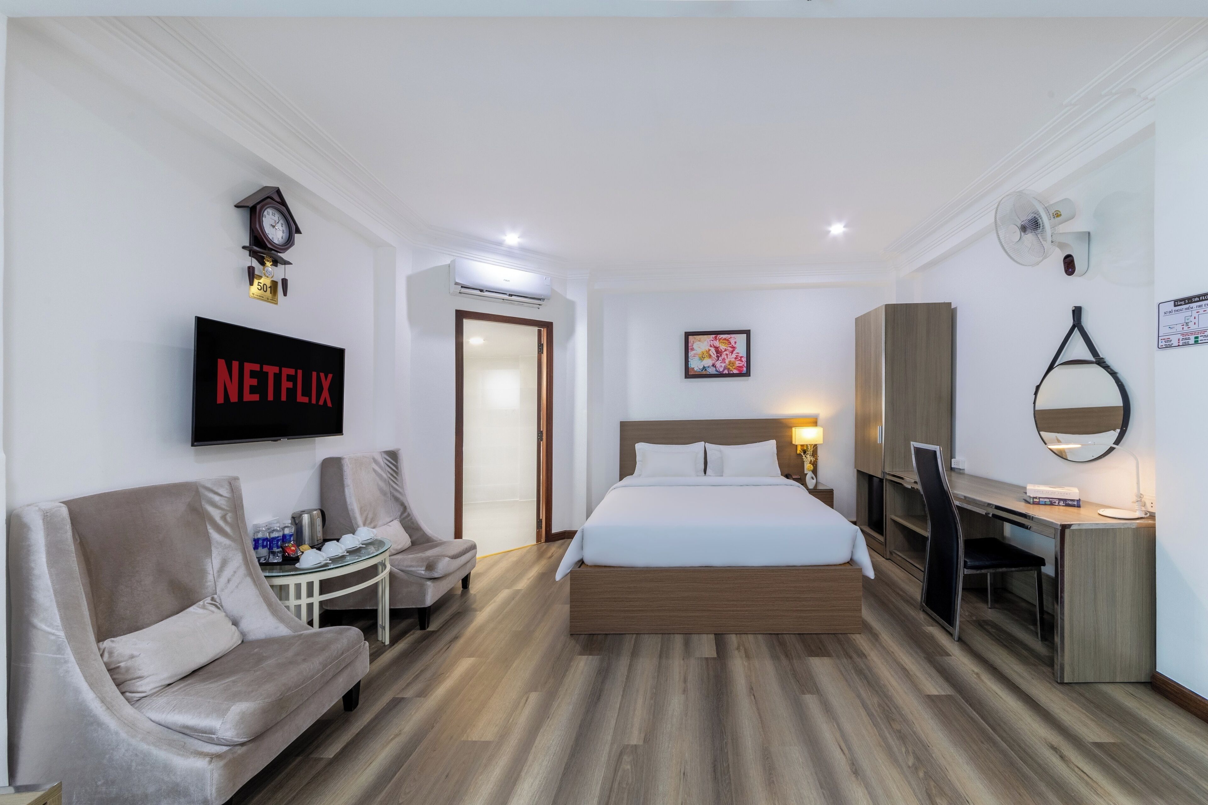 A modern hotel room with a bed, a desk, a chair, a TV displaying 'NETFLIX', a clock, an air conditioning unit, and a door.
