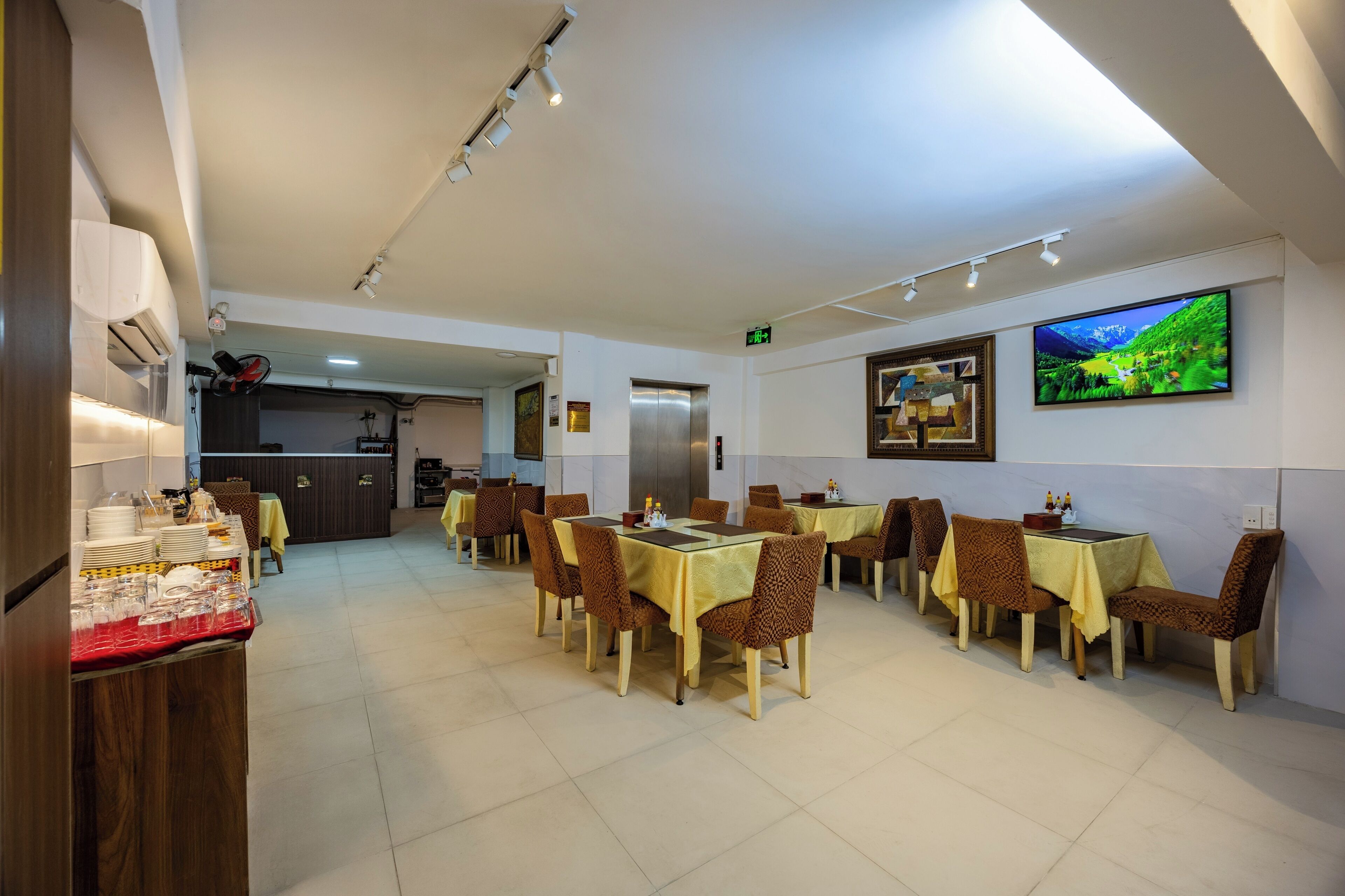 A dining area with tables and chairs, a kitchen in the background, and a television on the wall.
