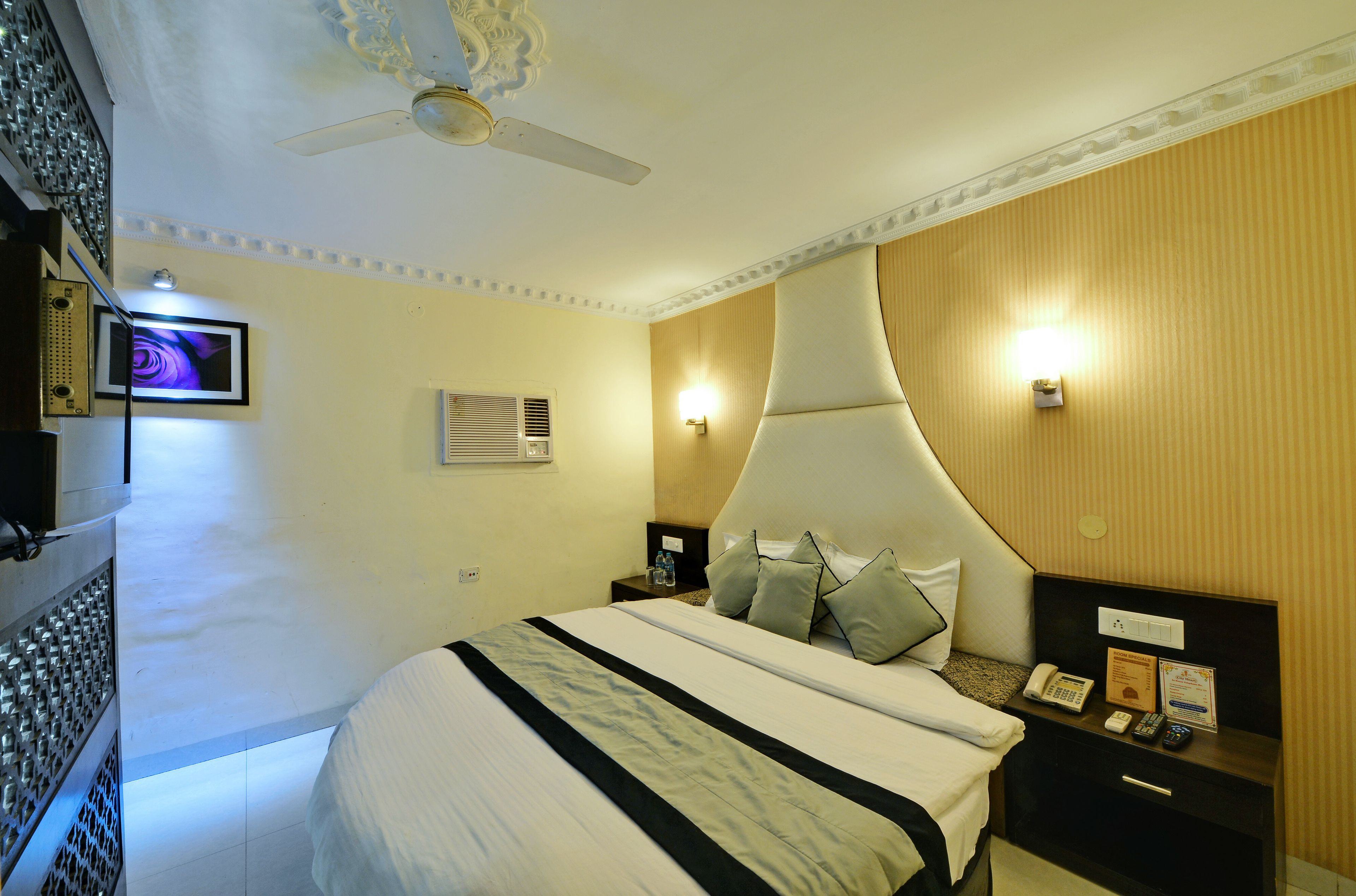 Habitación de hotel con una cama grande, una mesita de noche con teléfono y control remoto, un ventilador de techo y un televisor montado en la pared.