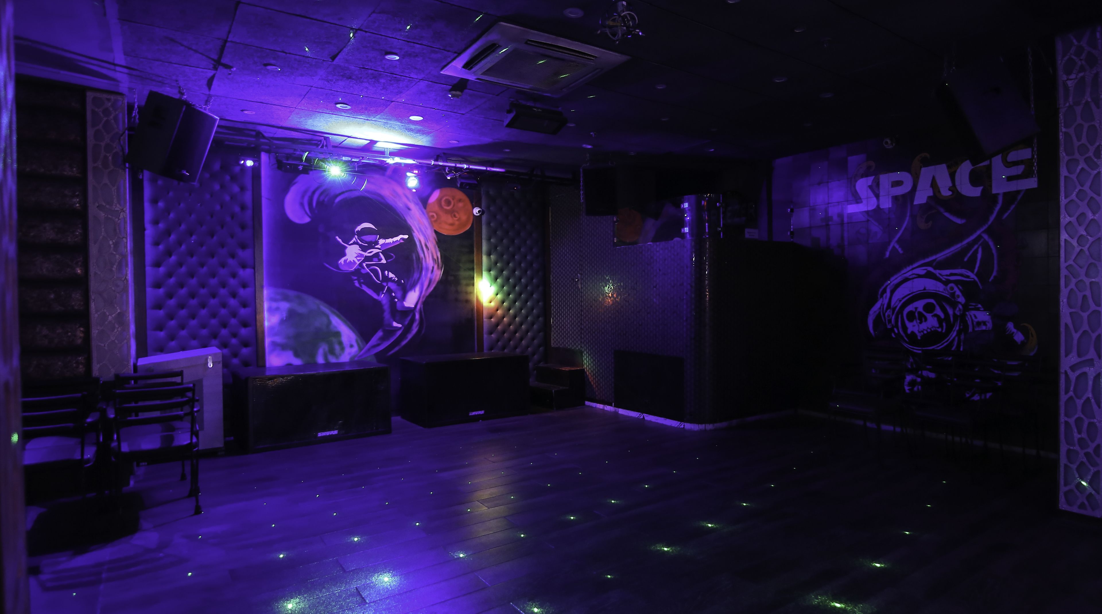 A room with a dark, moody ambiance, featuring a large wall mural of an astronaut, a plush seating area, and a wooden floor with embedded lights.