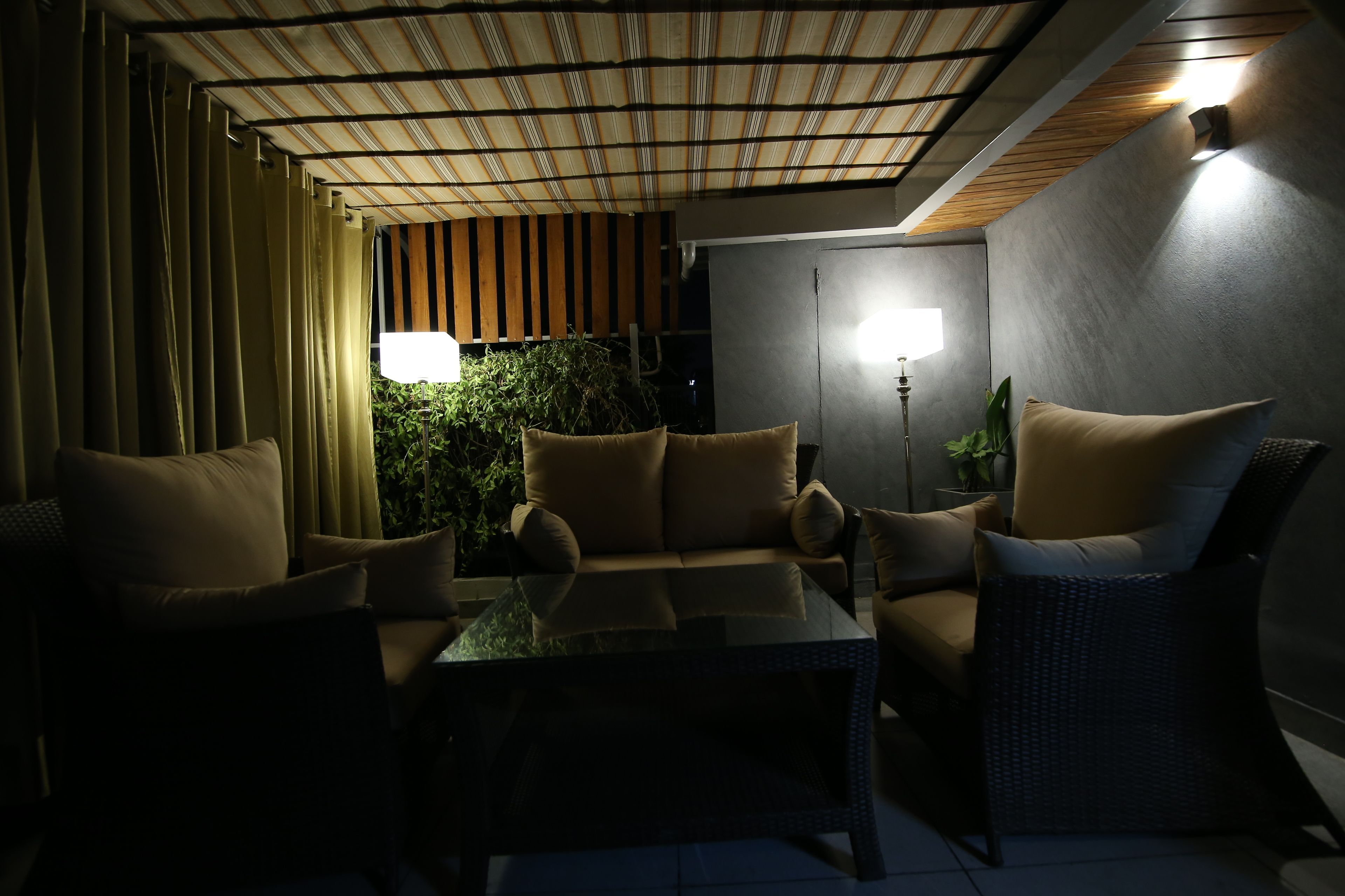 Un área de estar interior con poca luz, una mesa de vidrio, sillas acolchadas y una lámpara.