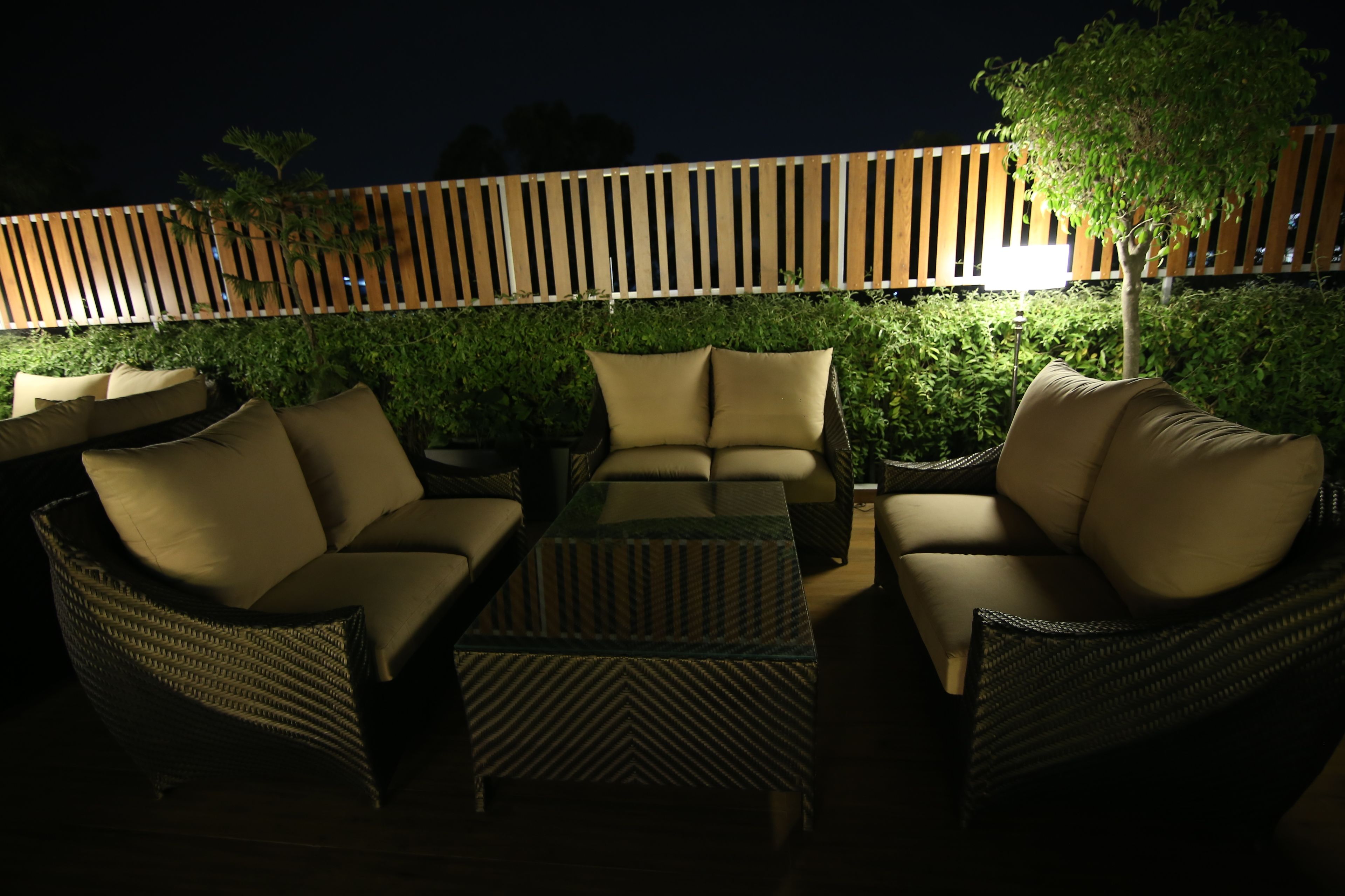 Un'area relax all'aperto con mobili in vimini e cuscini, una recinzione in legno e un siepe illuminata sullo sfondo.