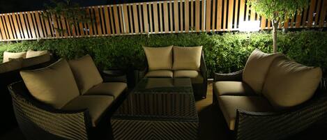 Un'area relax all'aperto con mobili in vimini e cuscini, una recinzione in legno e un siepe illuminata sullo sfondo.