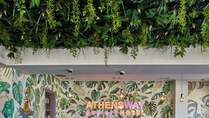 A reception area with a curved black counter, two wicker chairs, and a wall with a tropical leaf pattern. - Athens Way Pop Art Hotel (Athens)