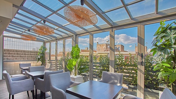A modern indoor space with glass walls, tables, and chairs, featuring a view of buildings and greenery outside. - Athens Way Pop Art Hotel (Athens)