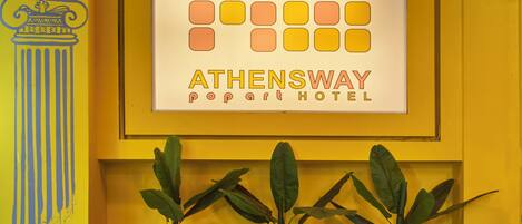 A reception area with a sign for Athenesway Popart Hotel, potted plants, and a decorative wall with a column illustration.