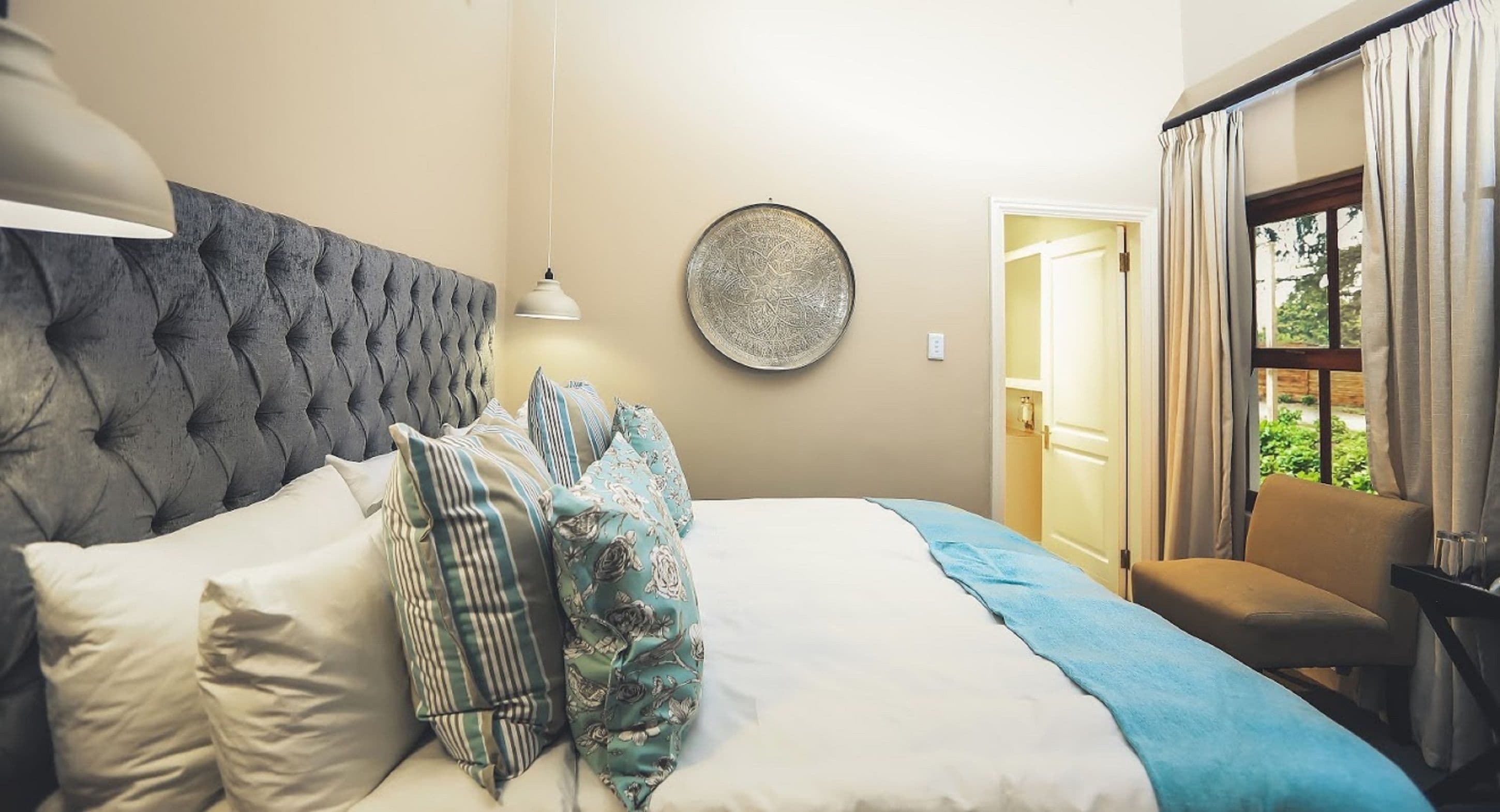 A bedroom with a tufted headboard, a round wall decoration, a bed with multiple pillows, a bedside table, and a chair.