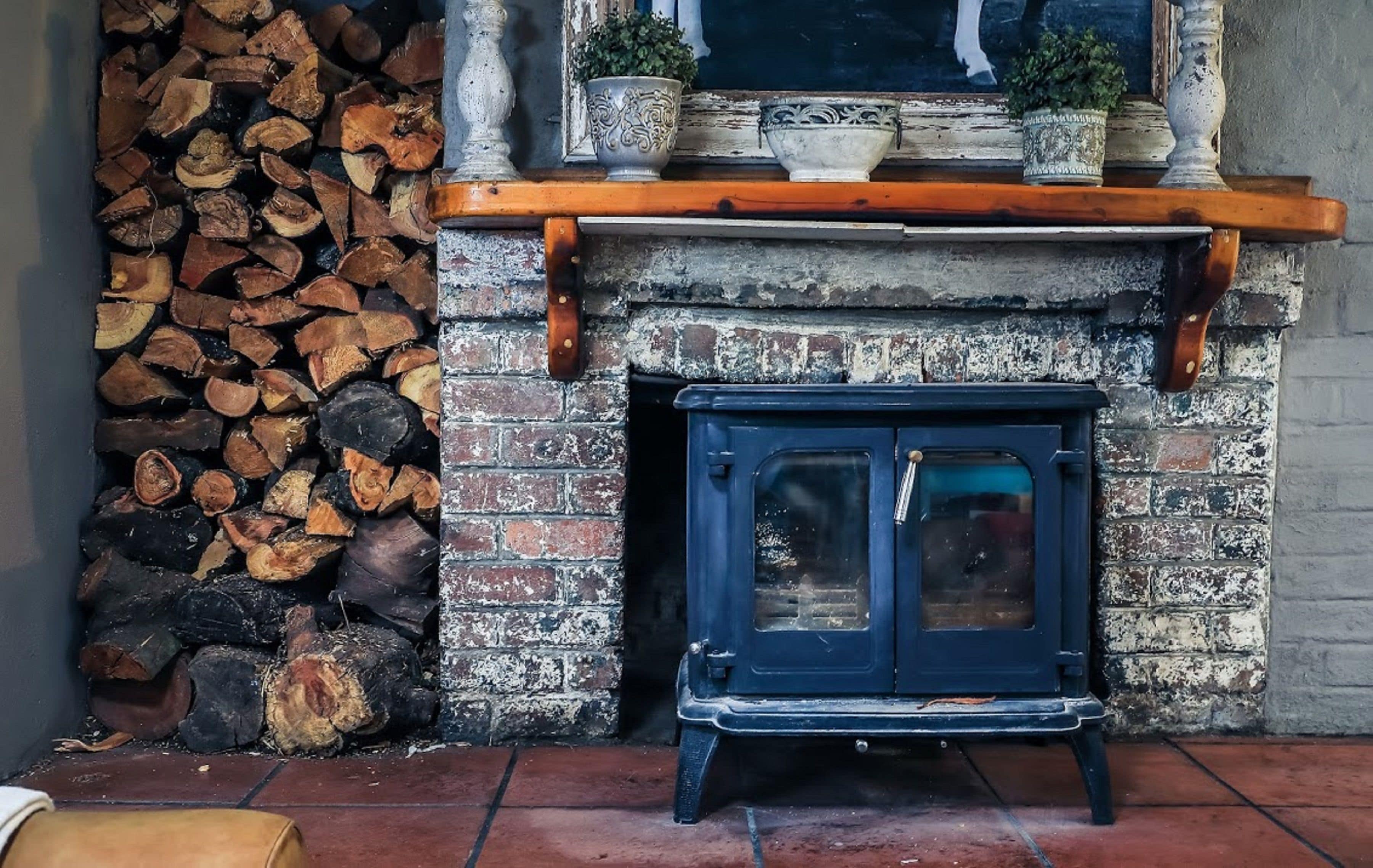A traditional fireplace with a stack of firewood to the left and a blue stove in front.