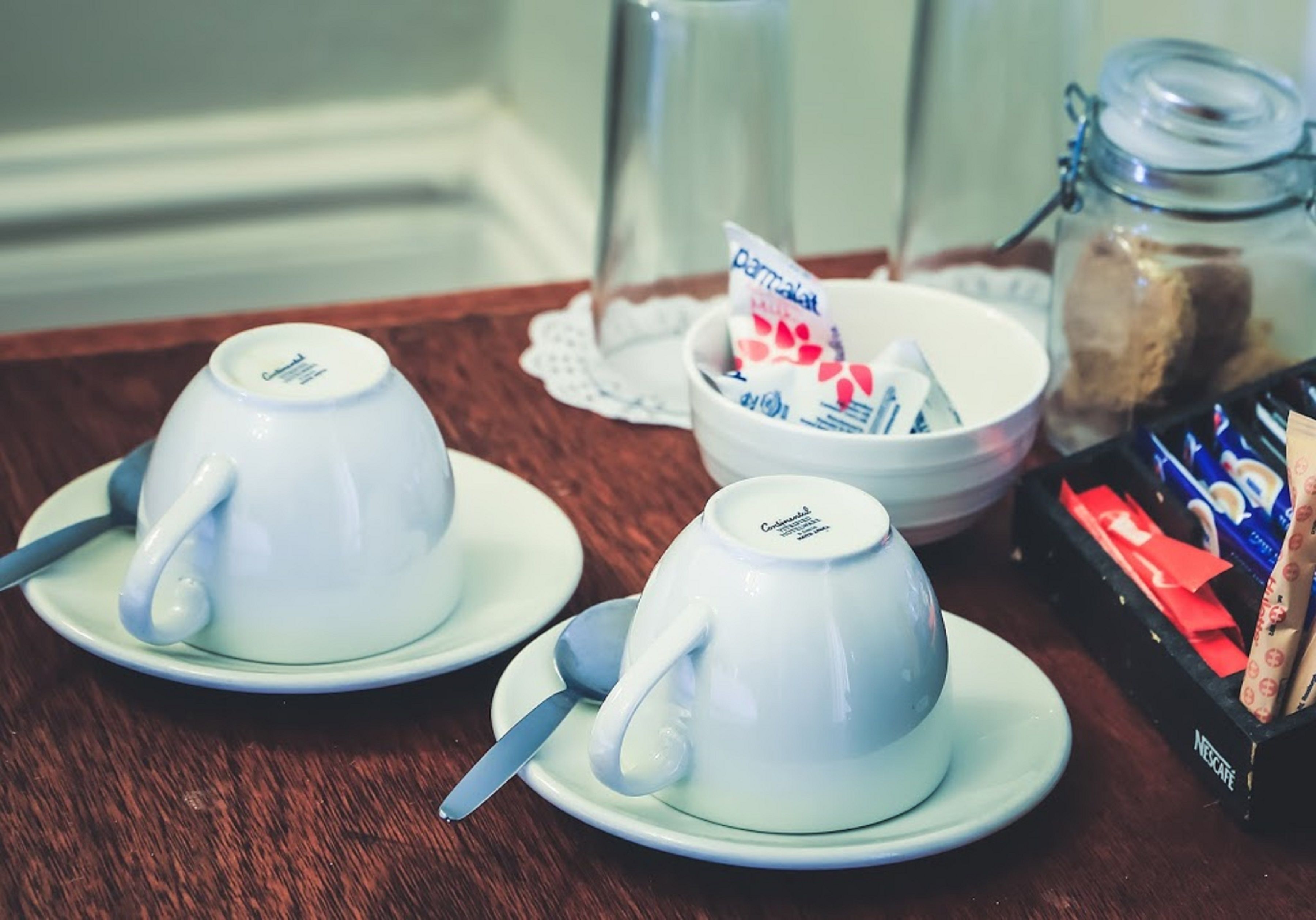Dos tazas blancas de cerámica sobre platillos, con cucharas, un tazón con un sobre de azúcar, un tarro de galletas y una caja de café Nescafe.