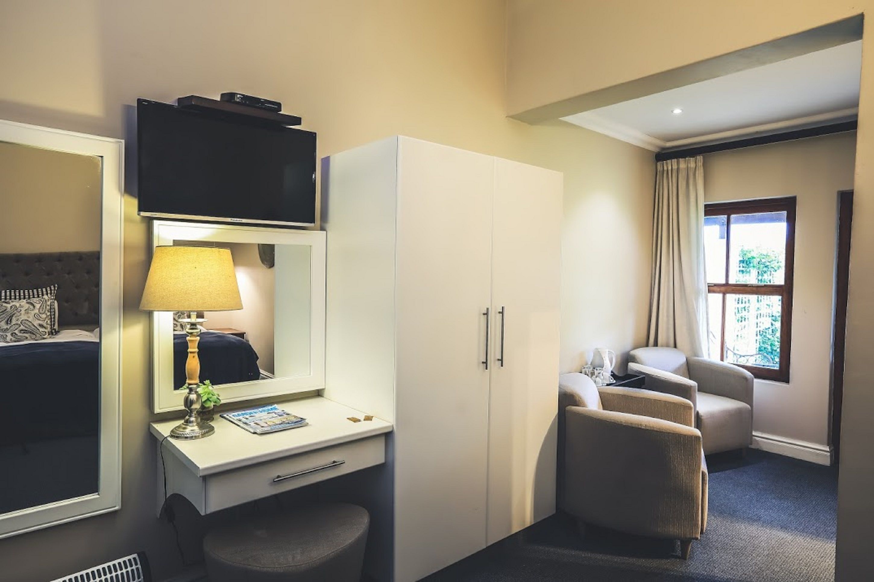 Une chambre d’hôtel comprenant un lit, un bureau avec une lampe, une télévision, une armoire, un coin salon avec deux fauteuils et une fenêtre avec des rideaux.