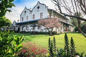 A large, white building with a balcony, surrounded by a well-maintained garden with pink flowers and green grass.