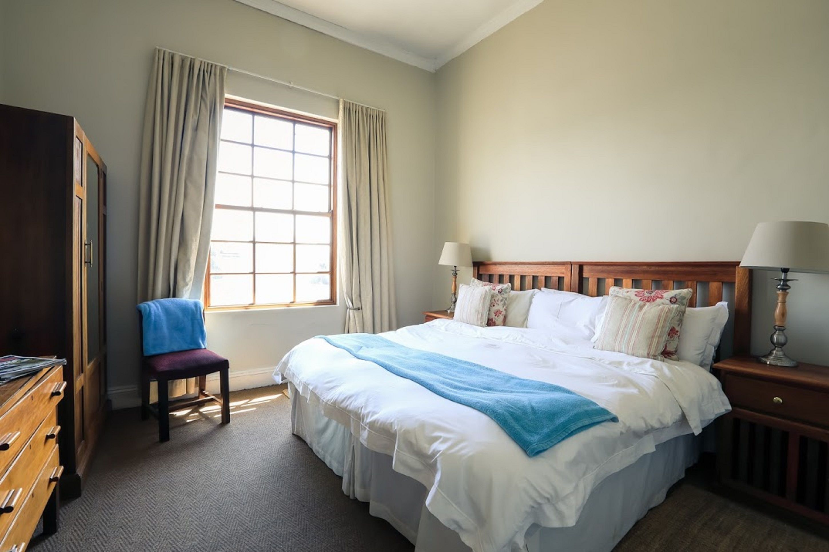 Un dormitorio ordenado con una cama grande, una silla, un aparador y una ventana con cortinas.