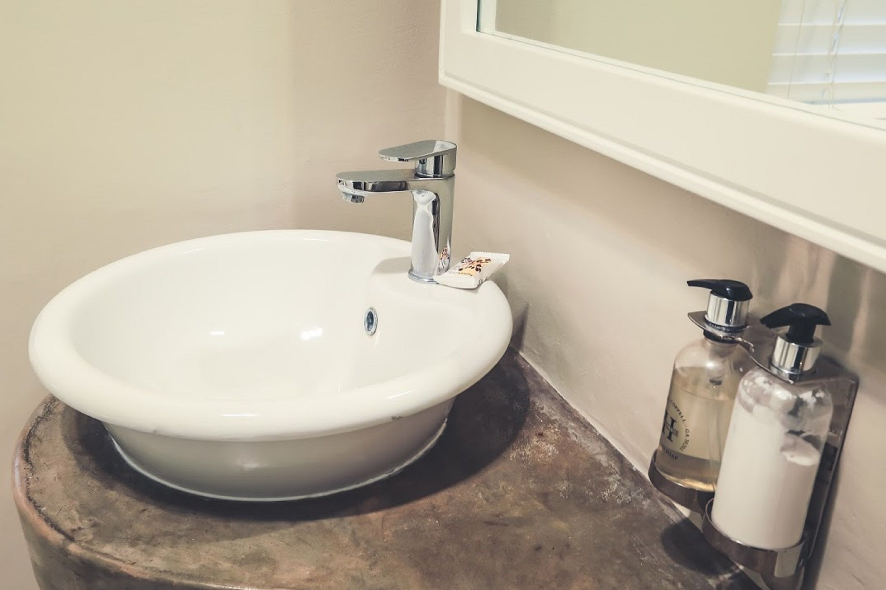 Un baño con un lavabo blanco, un espejo y dos dispensadores de jabón sobre un taburete de madera.