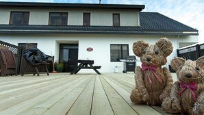 Um deck de madeira com churrasqueira, mesa de piquenique e dois ursos de palha decorando a fachada de um prédio com telhado e janelas pretas.