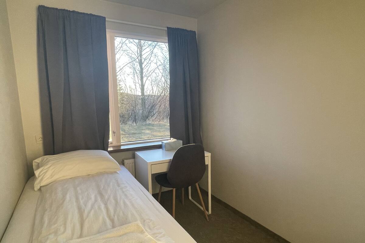 Habitación pequeña con cama individual, escritorio y silla, ventana con cortinas y radiador debajo del escritorio.