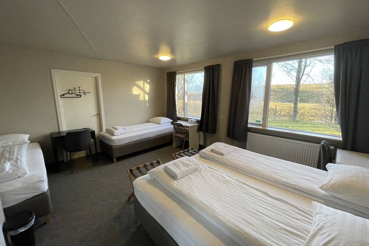 Habitación de hotel con dos camas, un escritorio, una silla, un radiador y una ventana con cortinas.