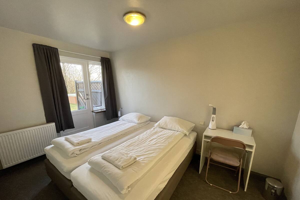 Una cama individual con ropa blanca, una mesita de noche con lámpara y una ventana con cortinas.