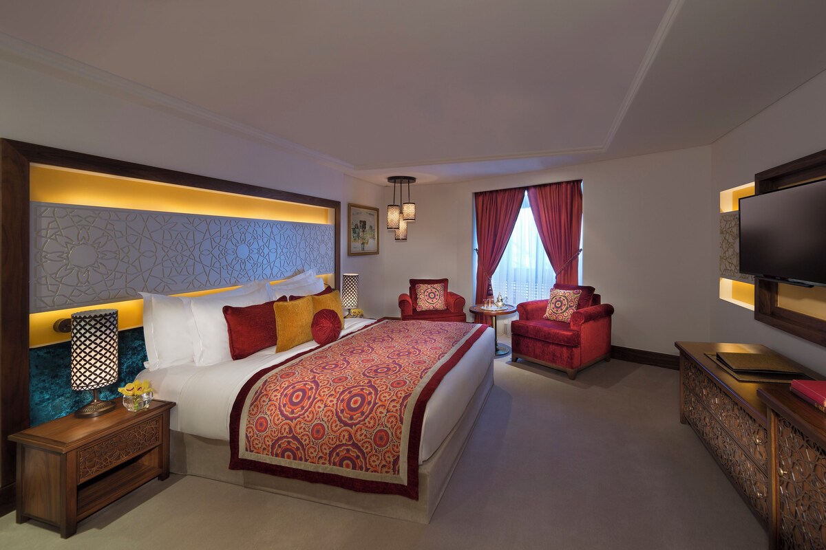 Habitación de hotel con una cama grande, dos sillones rojos, un televisor y un cabecero decorativo.