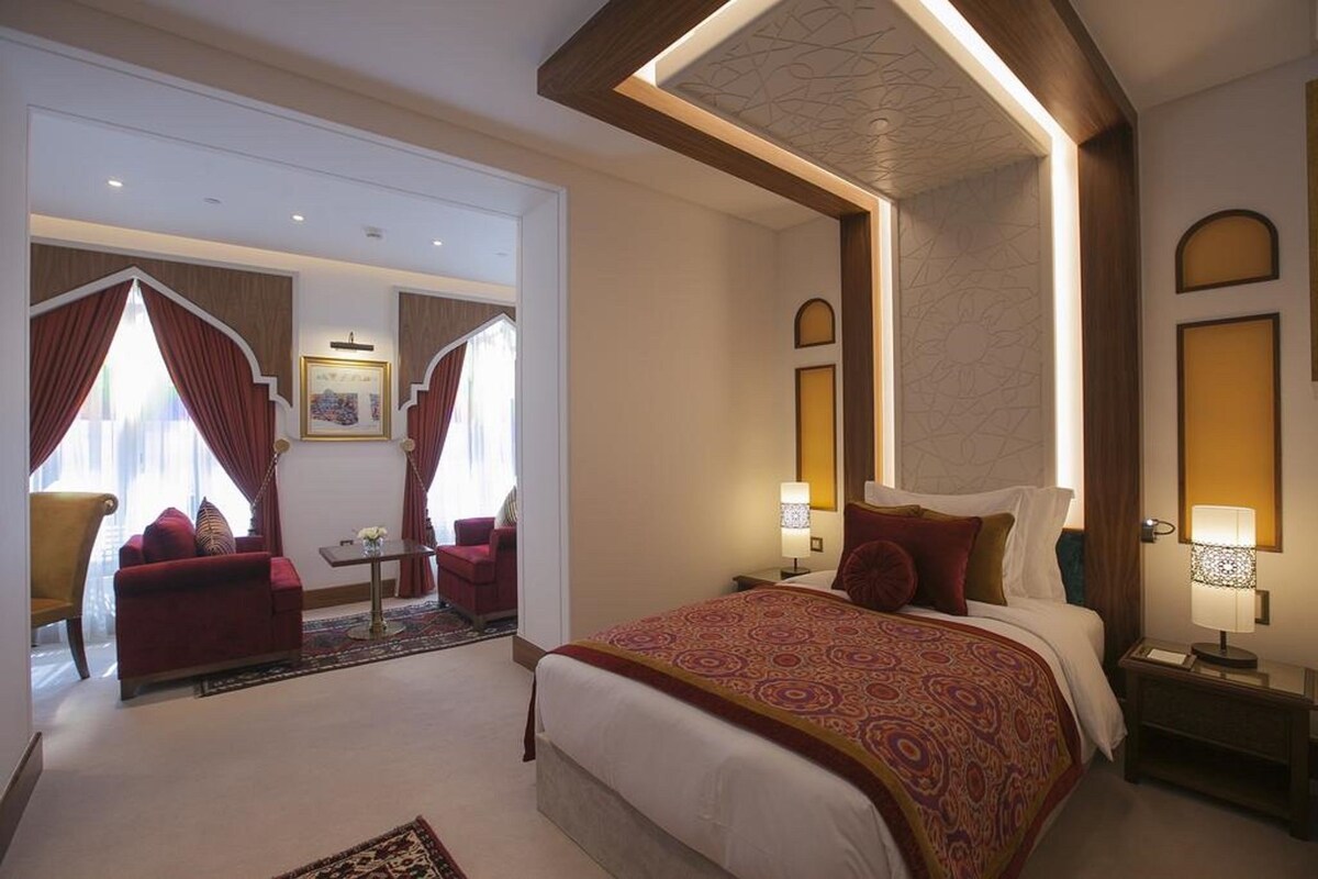 Habitación de hotel con una cama grande, un área de estar con un sofá y sillones, una mesita y vistas al exterior.